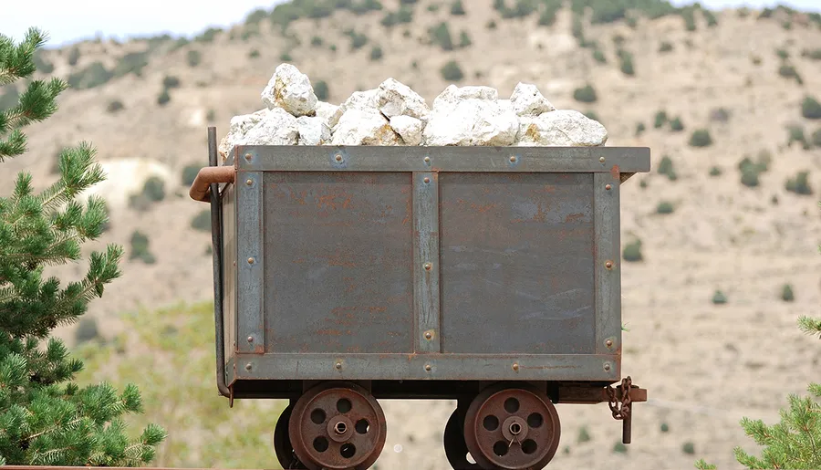 An old, rusted mining cart is filled with large rocks, set against a backdrop of hilly terrain and sparse vegetation.