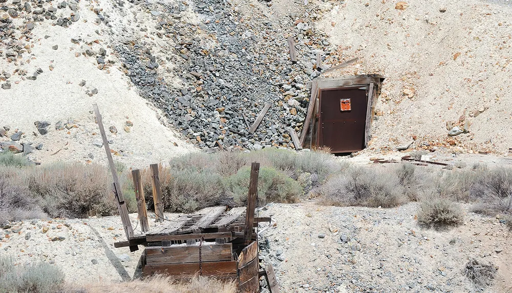The image shows a rustic metal door seemingly leading into a hillside with a collapsed wooden structure in the foreground set in a dry rocky landscape