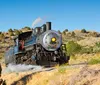 A vintage steam locomotive is chugging along a track through a dry hilly landscape under a clear blue sky