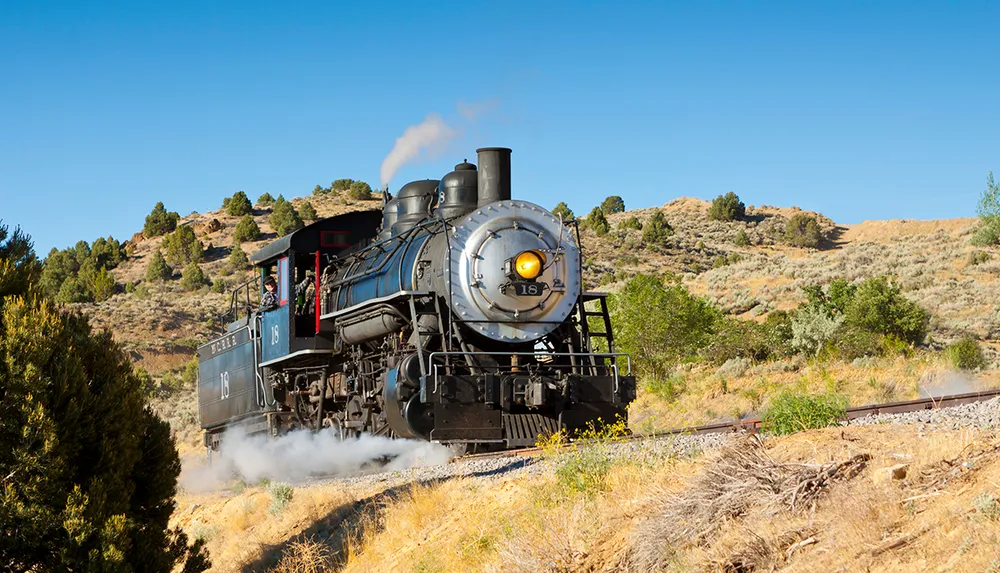 A vintage steam locomotive is chugging along a track through a dry hilly landscape under a clear blue sky