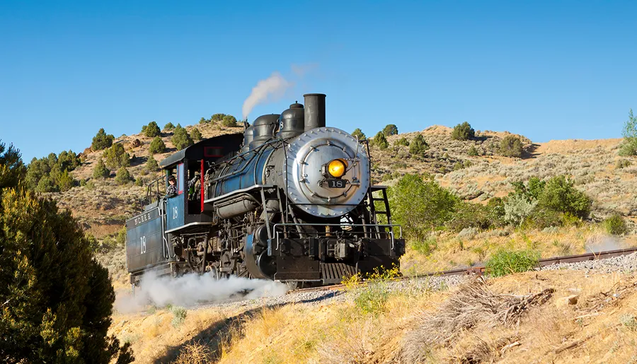 A vintage steam locomotive is chugging along a track through a dry, hilly landscape under a clear blue sky.