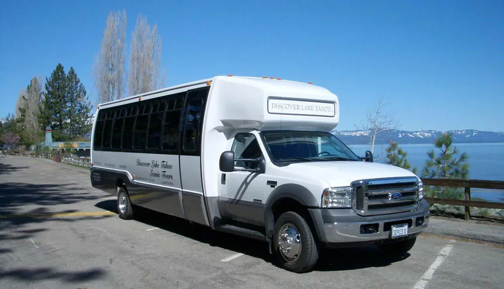 A white scenic tour bus with Discover Lake Tahoe written on its side is parked near a body of water with a mountainous landscape in the background