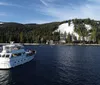 A white pleasure boat with people on deck is cruising on the water with a backdrop of pine trees and a cloudy sky