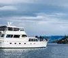 A white pleasure boat with people on deck is cruising on the water with a backdrop of pine trees and a cloudy sky