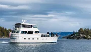 A white pleasure boat with people on deck is cruising on the water with a backdrop of pine trees and a cloudy sky.