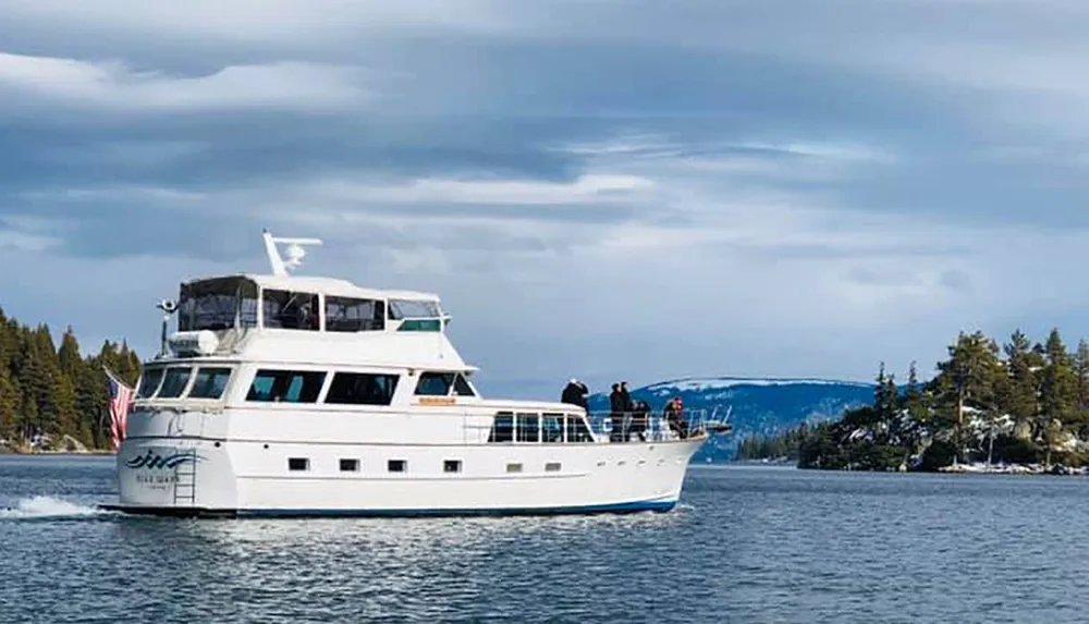 A white pleasure boat with people on deck is cruising on the water with a backdrop of pine trees and a cloudy sky