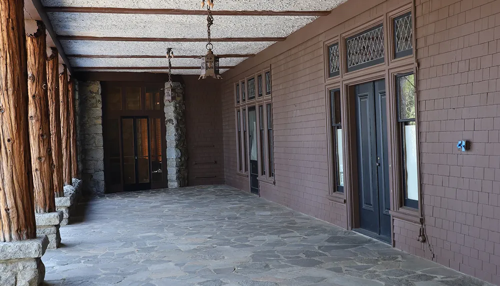 The image shows an outdoor covered passageway with rustic wood and stone columns a tiled floor and a series of doors and windows along a brick wall