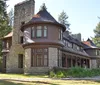 This is a photo of a stately historic house with stone foundations and a round turret surrounded by mature trees under a clear blue sky