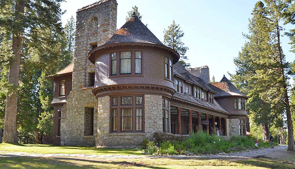 This is a photo of a stately historic house with stone foundations and a round turret surrounded by mature trees under a clear blue sky