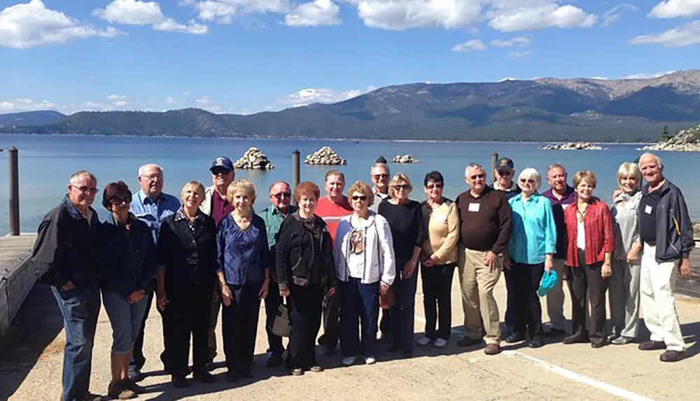 A group of mature adults poses for a photo on a pier with a mountainous lakeside backdrop under a clear sky