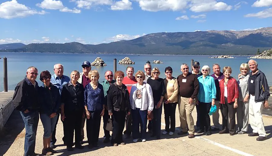 A group of mature adults poses for a photo on a pier with a mountainous lakeside backdrop under a clear sky.