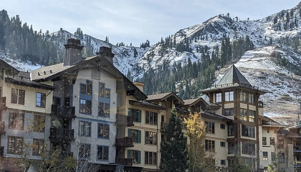 The image shows alpine-style buildings nestled at the foot of a lightly snow-covered mountain