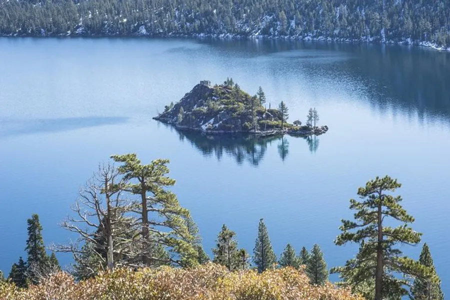 The image features a small, verdant island covered with trees, situated in the middle of a calm, blue lake, with surrounding coniferous forest and a backdrop of mountain slopes.