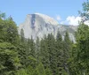A person is sitting on a log by a tranquil river gazing at the impressive granite cliffs and lush greenery of a mountainous landscape