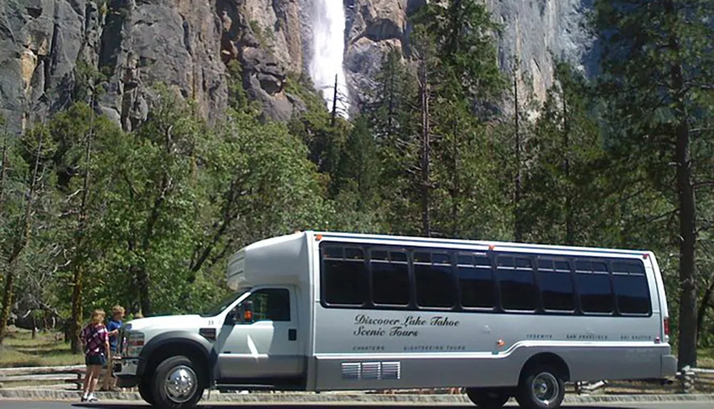 A tour bus with the text Discover Lake Tahoe Scenic Tours is parked with passengers boarding or disembarking in front of a towering waterfall amidst a forested landscape