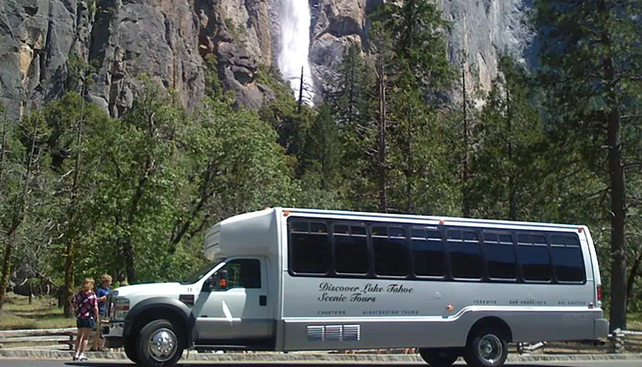 A tour bus with the text Discover Lake Tahoe Scenic Tours is parked with passengers boarding or disembarking in front of a towering waterfall amidst a forested landscape.
