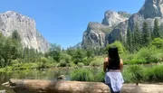 A person is sitting on a log by a tranquil river, gazing at the impressive granite cliffs and lush greenery of a mountainous landscape.