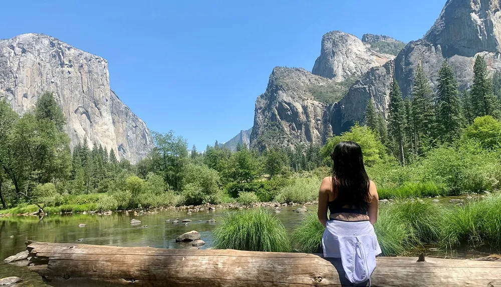 A person is sitting on a log by a tranquil river gazing at the impressive granite cliffs and lush greenery of a mountainous landscape