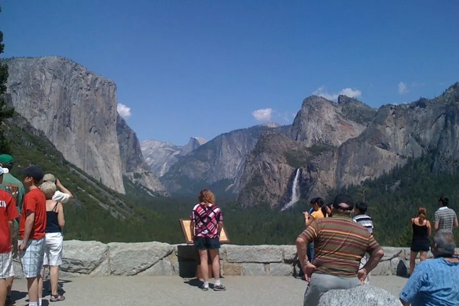 Visitors are admiring the scenic view of a valley with a waterfall and towering cliffs under a clear blue sky.