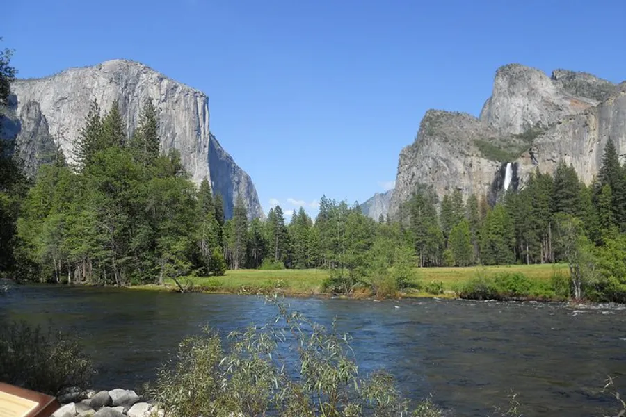 The image showcases a serene river flowing through a lush meadow with towering granite cliffs and a waterfall in the distance, likely a scene from Yosemite National Park.