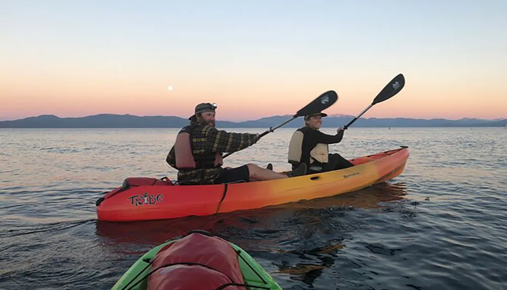 Two people in a tandem kayak are paddling on calm waters with the sunset and a full moon in the background