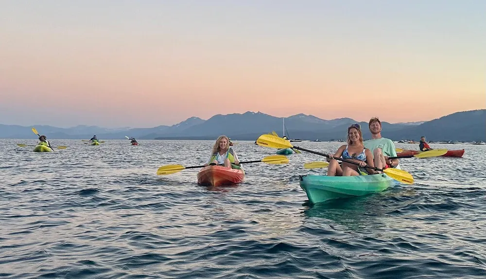 A group of people are joyfully kayaking on a calm body of water against a backdrop of a mountainous horizon at sunset