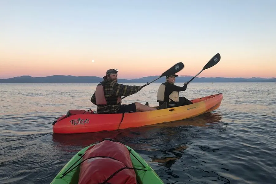 Two people are paddling a tandem kayak on tranquil waters at sunset, with a beautiful sky gradient in the background and a view of the tops of other kayaks in the foreground.