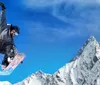 A snowboarder in colorful winter gear is performing a trick on a sunny mountain slope with clear blue skies in the background