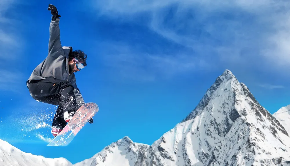 A snowboarder is performing a mid-air trick against a backdrop of clear blue skies and snowy mountains
