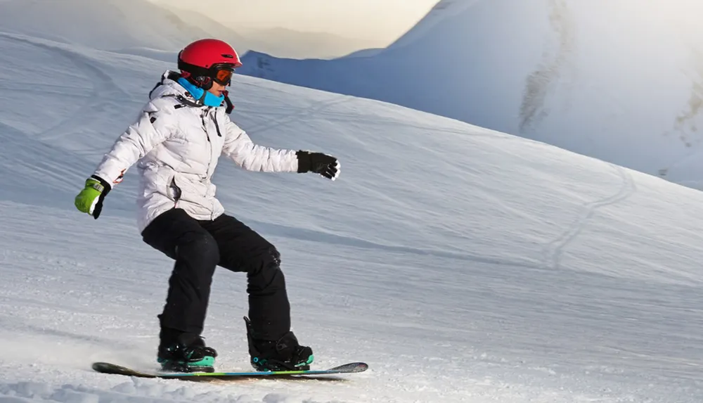 A snowboarder in a white jacket and helmet glides down a snowy slope with mountains in the background