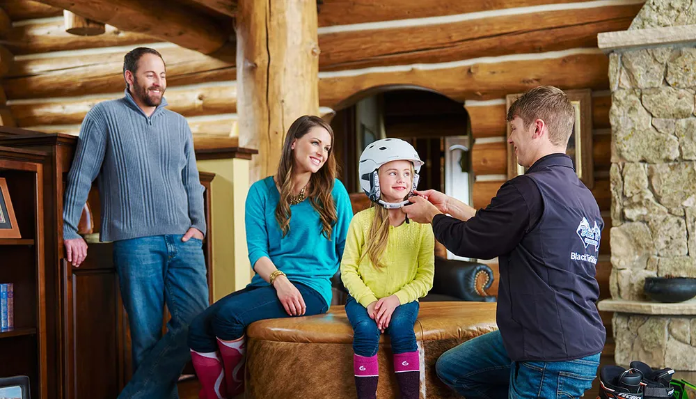 A man is fitting a helmet on a young girl while two adults watch with smiles in a cozy rustic interior setting