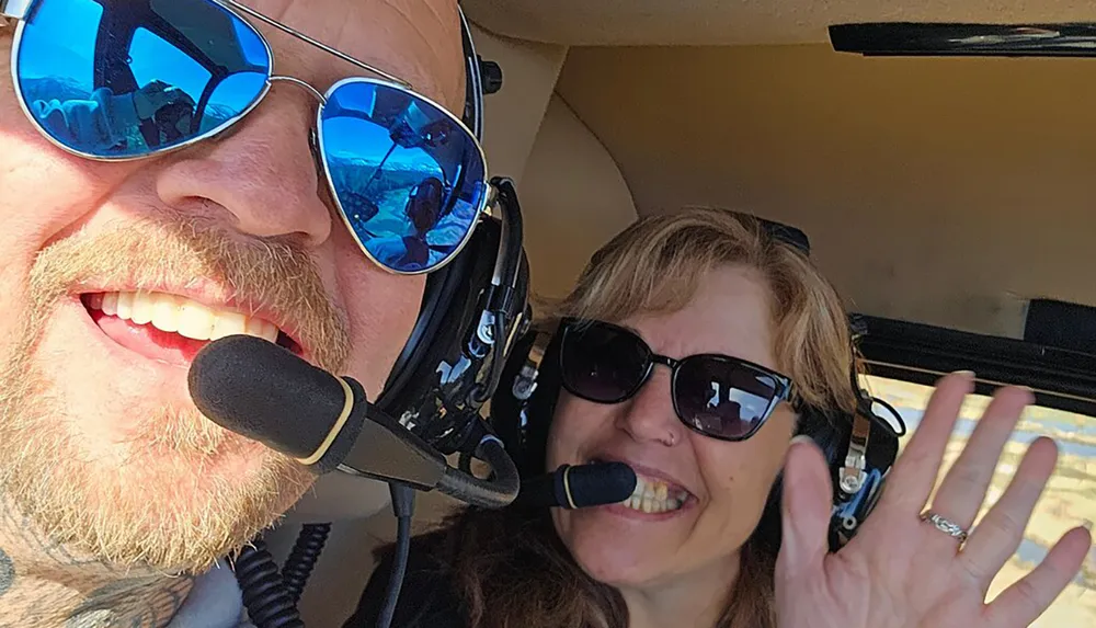 A man and a woman wearing sunglasses and headsets with microphones are smiling and waving inside an aircraft cockpit