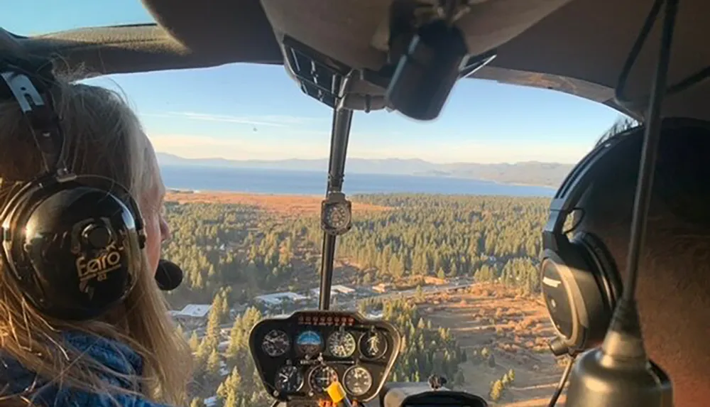 Two people wearing headsets are inside a helicopter flying over a landscape with trees and a view of a lake in the distance