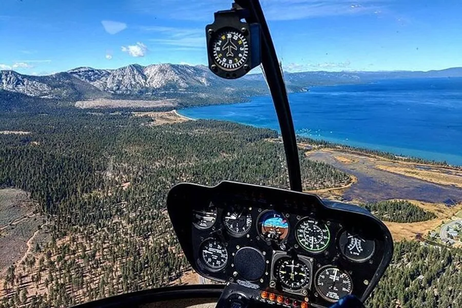 The image shows the cockpit view from a helicopter flying over a scenic landscape with a large lake and mountainous terrain in the background.