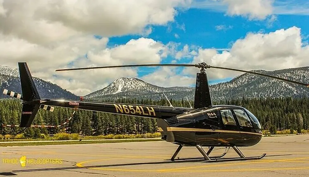 A helicopter is parked on a landing pad with snow-capped mountains and a partly cloudy sky in the background