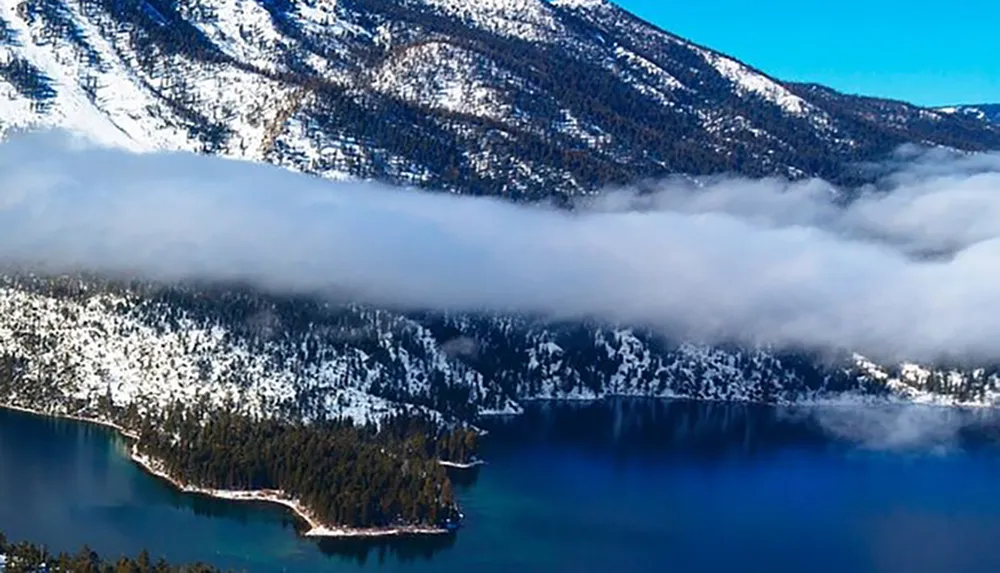 A thick blanket of low-lying clouds nestles in a mountainous snow-covered landscape above a deep blue lake