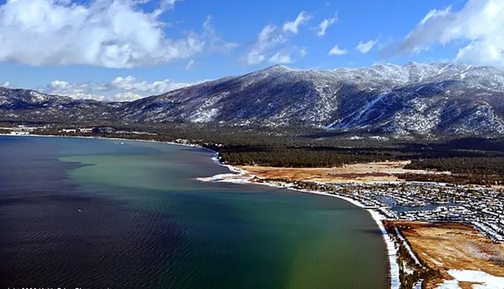 An aerial view of a snowy mountain range beside a large body of water with a beachfront and a partially snow-covered landscape