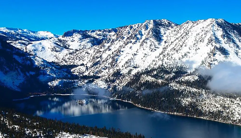 A serene winter landscape showcases a snow-covered mountainous terrain with a crystal-clear lake nestled amidst the peaks under a bright blue sky