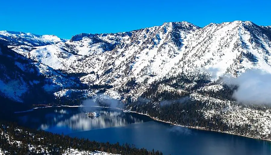 A serene winter landscape showcases a snow-covered mountainous terrain with a crystal-clear lake nestled amidst the peaks under a bright blue sky.