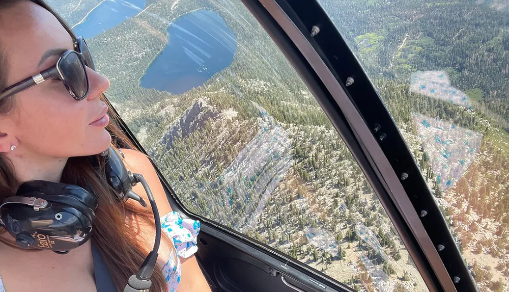 A person wearing sunglasses and a headset is looking out the window of a helicopter at a scenic landscape below