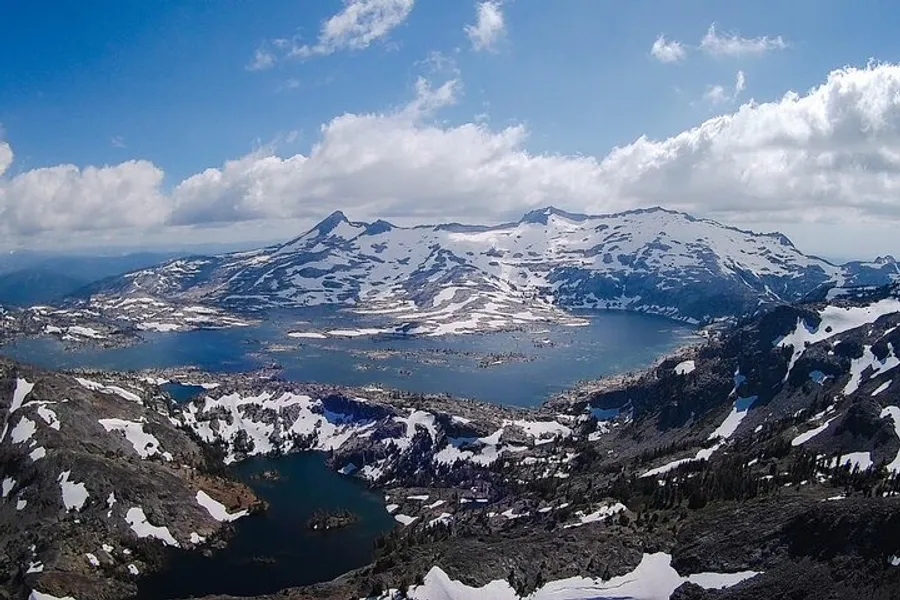 The image showcases a panoramic alpine landscape featuring snow-capped mountains and a series of lakes nestled among the rugged terrain under a partly cloudy sky.