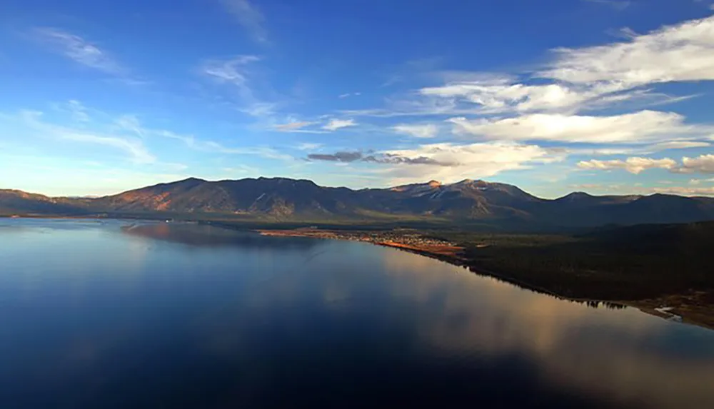 A serene lake reflects the sky surrounded by a lush forest and a mountain range under a partly cloudy sky