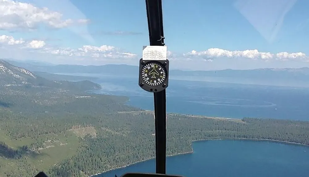 The image shows a breathtaking aerial view of a large lake and forested area through the windshield of an aircraft with the aircrafts compass positioned centrally in the frame
