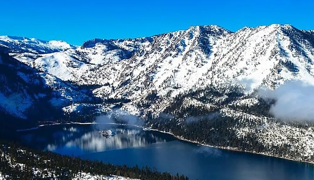 The image features a serene snow-capped mountain range overlooking a forested lakeside with patches of clouds draped around the scenery