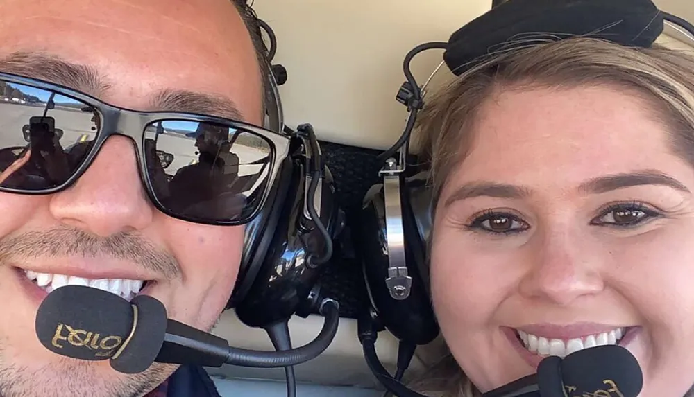 Two smiling people wearing aviation headsets are taking a close-up selfie with reflective sunglasses revealing someones hand holding the camera