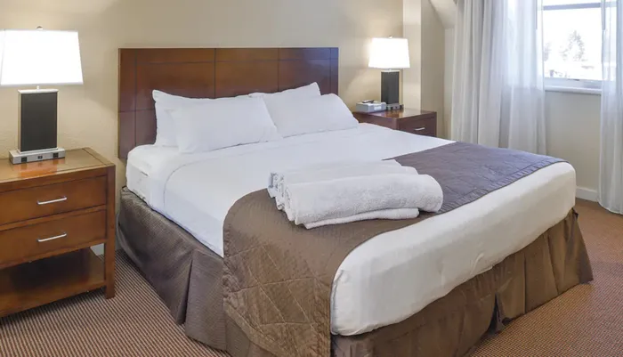 The image shows a neatly made hotel room bed with white linens and a brown bedspread flanked by matching nightstands with lamps