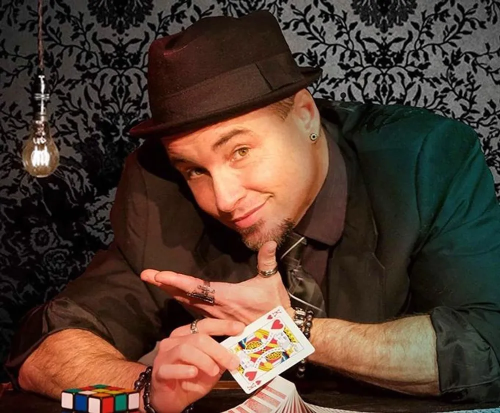 A person in a fedora and dark shirt is smiling at the camera pointing to a fanned-out hand of playing cards with a Rubiks cube and a clear light bulb visible nearby set against a patterned backdrop