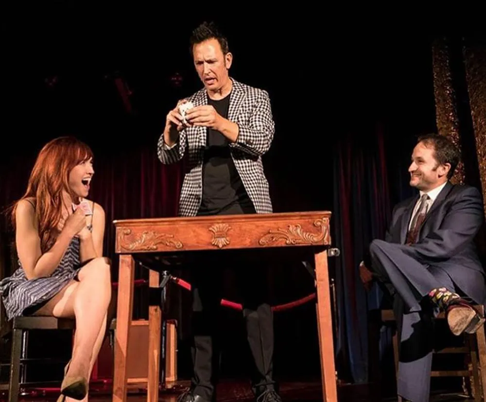 A magician performs a trick on stage with a deck of cards that delights a woman seated to the left and amuses a man in a suit seated to the right