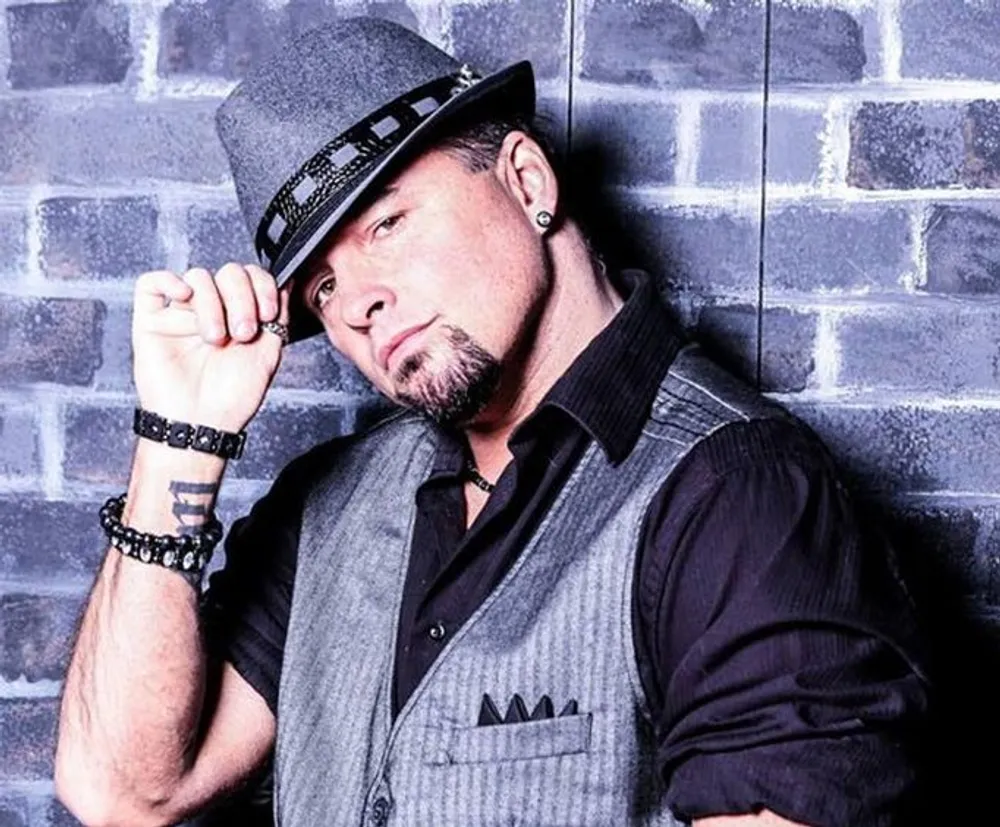 A person is posing with a confident attitude against a brick wall wearing a fedora a vest and accessorized with beaded bracelets