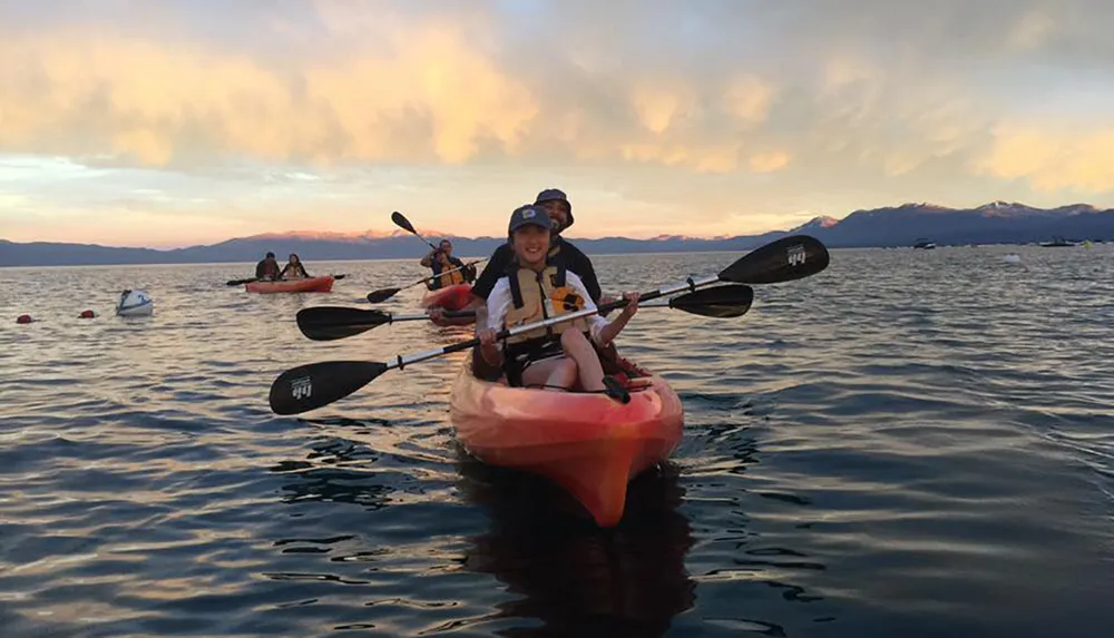 A group of people are kayaking on calm waters with a backdrop of a beautiful sunset and mountains